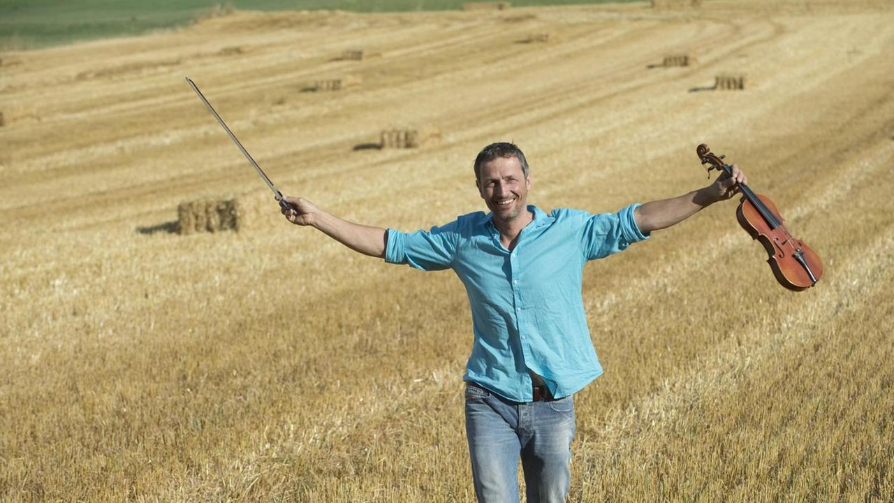 Ein Geiger läuft in Jeans und Leinenhemd mit ausgestreckten Armen über ein gemähtes Feld. Ein Geiger läuft in Jeans und Leinenhemd mit ausgestreckten Armen über ein gemähtes Feld.