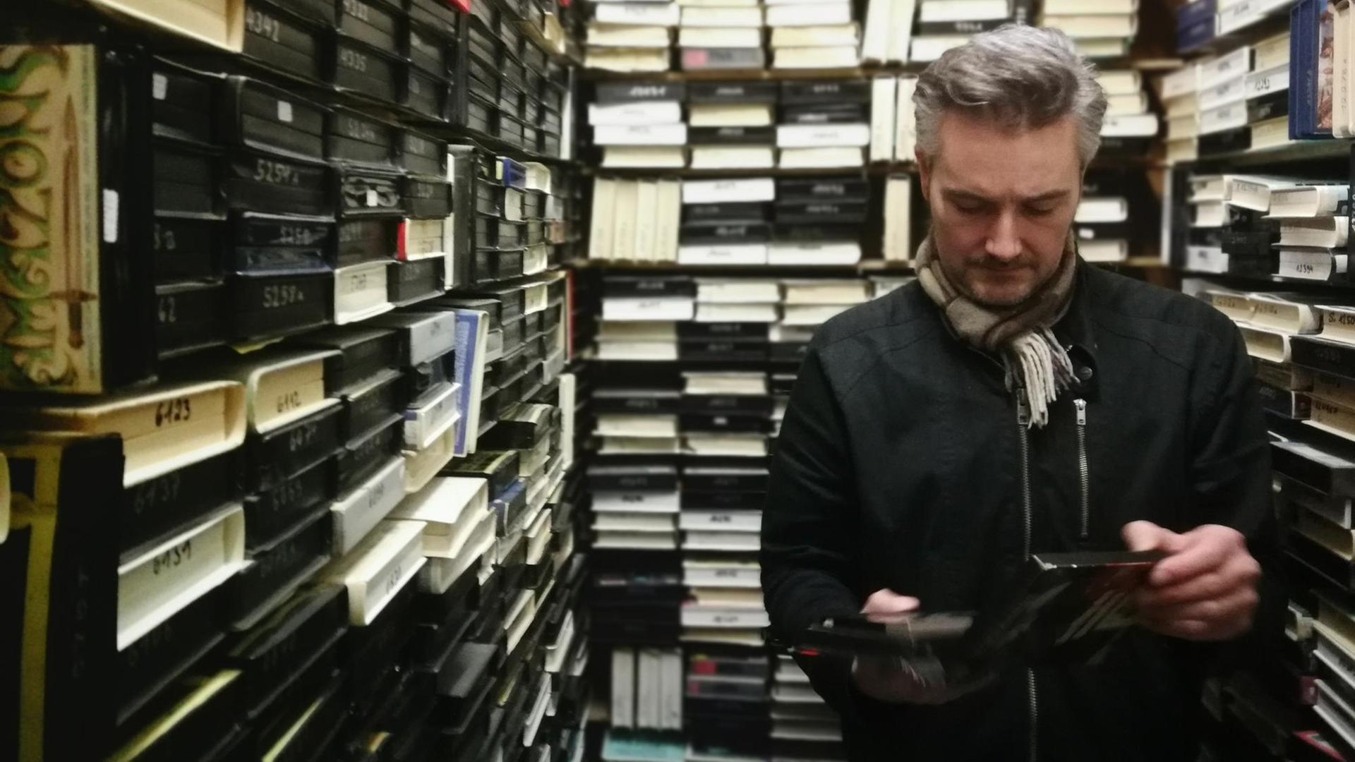 Ein Mann steht zwischen Regalen voller Videokassetten. Ein Mann steht zwischen Regalen voller Videokassetten.