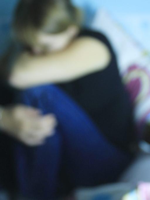 Eine verzweifelte junge Frau hockt auf einem Bett. Im Vordergrund: Tabletten.