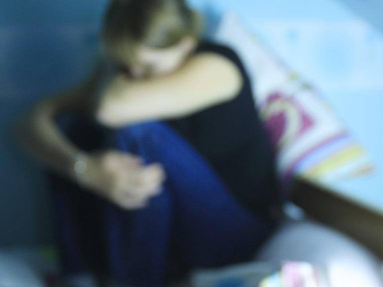 Eine verzweifelte junge Frau hockt auf einem Bett. Im Vordergrund: Tabletten.