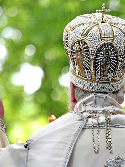 Die Rückansicht eines Griechisch-orthodoxen Priesters in der Türkei. Er breitet die Hände zu einem Segen aus und trägt eine bestickte weisse Robe und eine reich verzierte Bischofskrone.