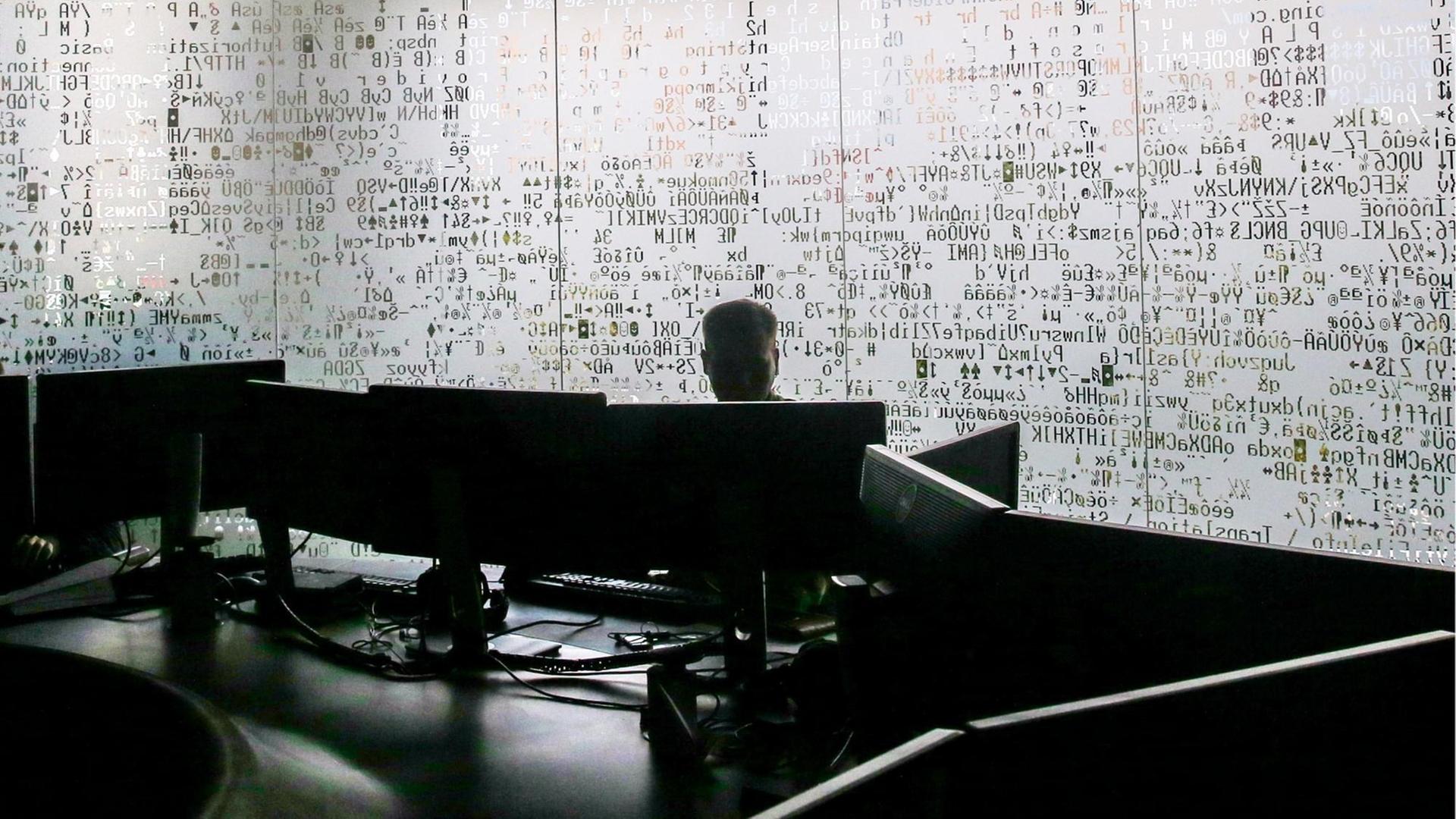 Die verschattete Silhouette eines Mannes an einem Schreibtisch, im Hintergrund eine Milchglaswand mit Code zum Teil in kyrillischen Buchstaben. Die verschattete Silhouette eines Mannes an einem Schreibtisch, im Hintergrund eine Milchglaswand mit Code zum Teil in kyrillischen Buchstaben.