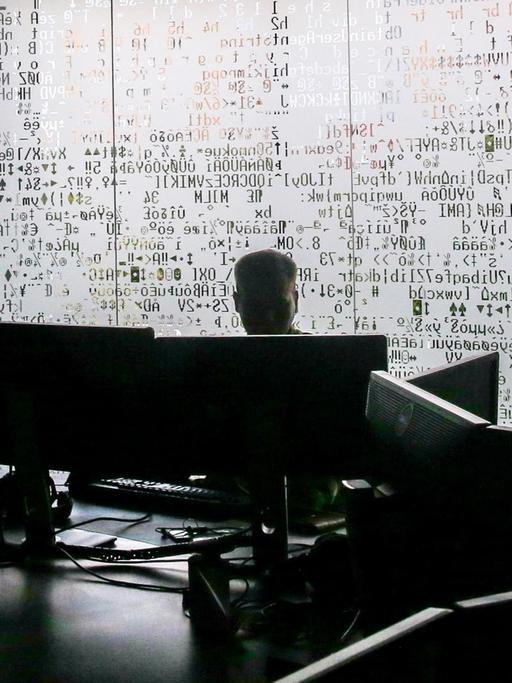 Die verschattete Silhouette eines Mannes an einem Schreibtisch, im Hintergrund eine Milchglaswand mit Code zum Teil in kyrillischen Buchstaben.