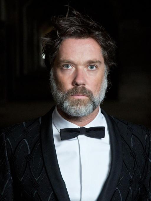 Der Musiker Rufus Wainwright steht in schwarzem Anzug mit Fliege vor einem schwarzen Hintergrund