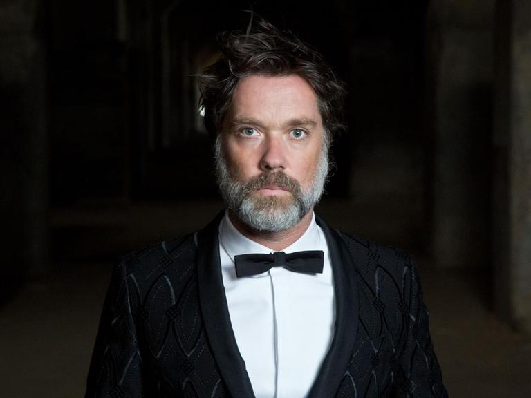 Der Musiker Rufus Wainwright steht in schwarzem Anzug mit Fliege vor einem schwarzen Hintergrund