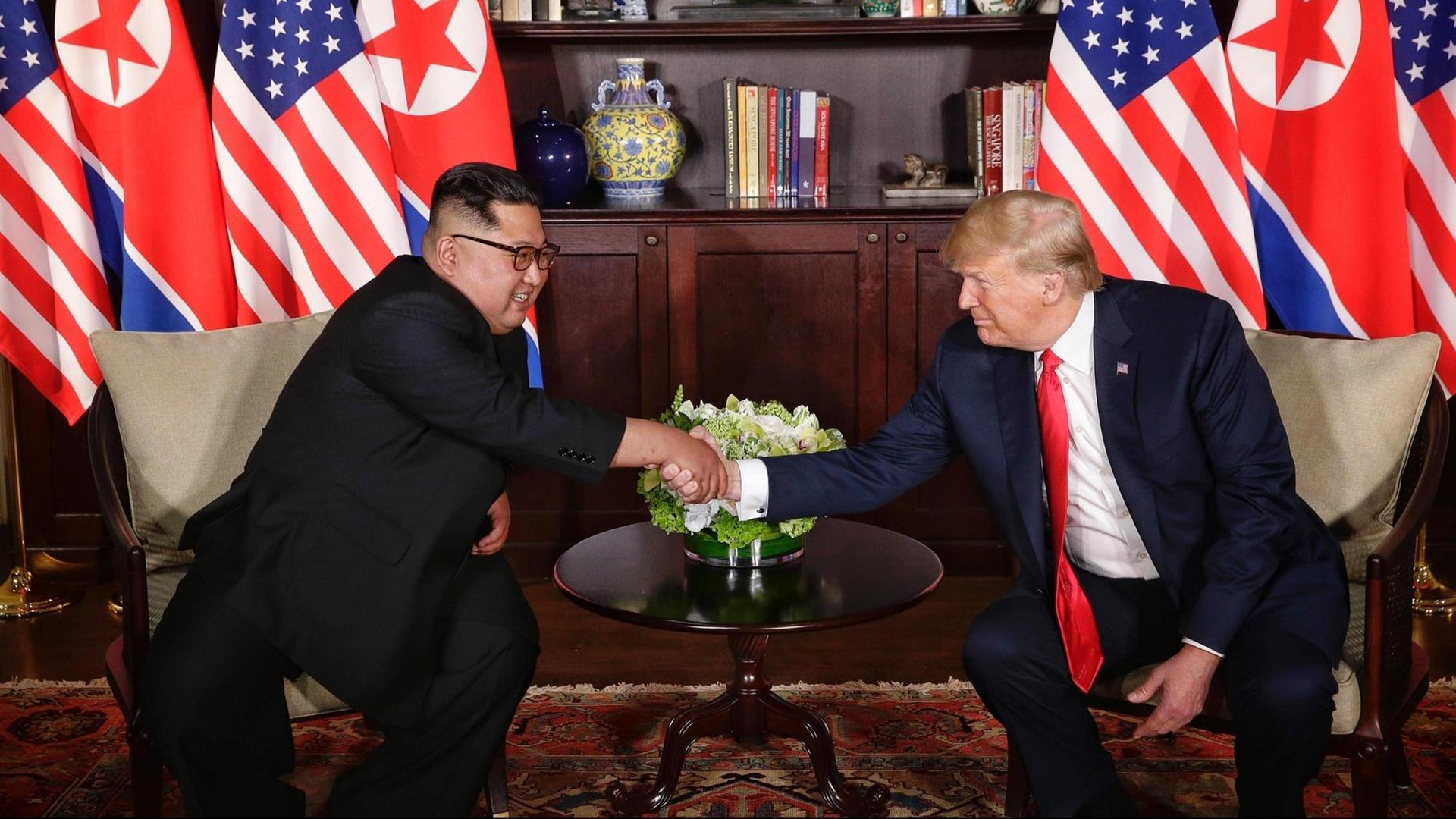 Kim (l.) und Trump sitzen nebeneinander an einem kleinen runden Tisch und reichen sich die Hand. Hinter ihm die Nationalflaggen. Kim (l.) und Trump sitzen nebeneinander an einem kleinen runden Tisch und reichen sich die Hand. Hinter ihm die Nationalflaggen.