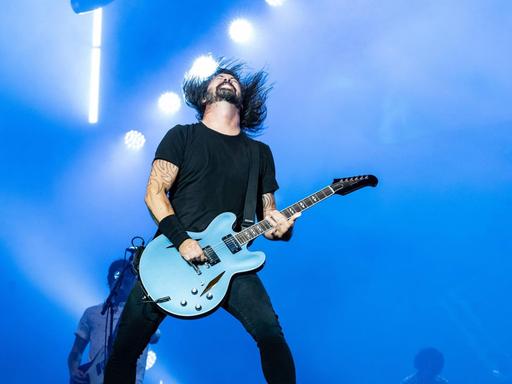 Dave Grohl spielt bei einem Konzert mit den Foo Fighters auf der Bühne Gitarre und wirft seinen Kopf mit den langen Haaren nach hinten.