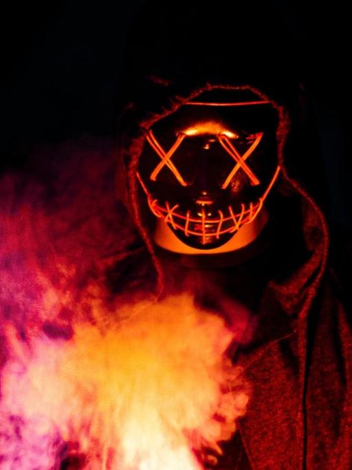 Symbolbild für Dämonen: eine Gestalt mit gruselige Maske unter einer Kapuze mit Feuer in der Hand.