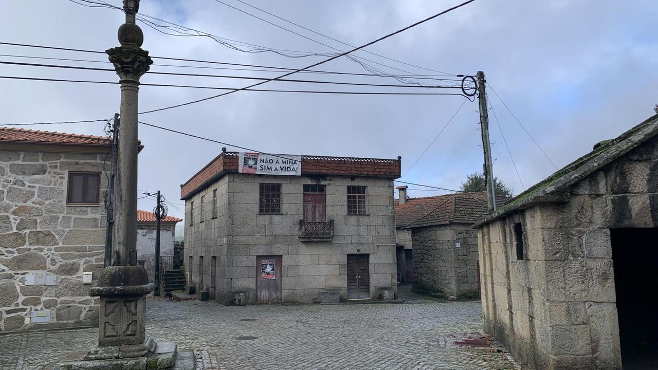 Ein kleiner Platz, der eingerahmt wird von drei alten quadratischen Steinhäusern. An einem der Häuser hängt direkt unter dem Dach ein weißes rechteckiges Plakat. Ein kleiner Platz, der eingerahmt wird von drei alten quadratischen Steinhäusern. An einem der Häuser hängt direkt unter dem Dach ein weißes rechteckiges Plakat.
