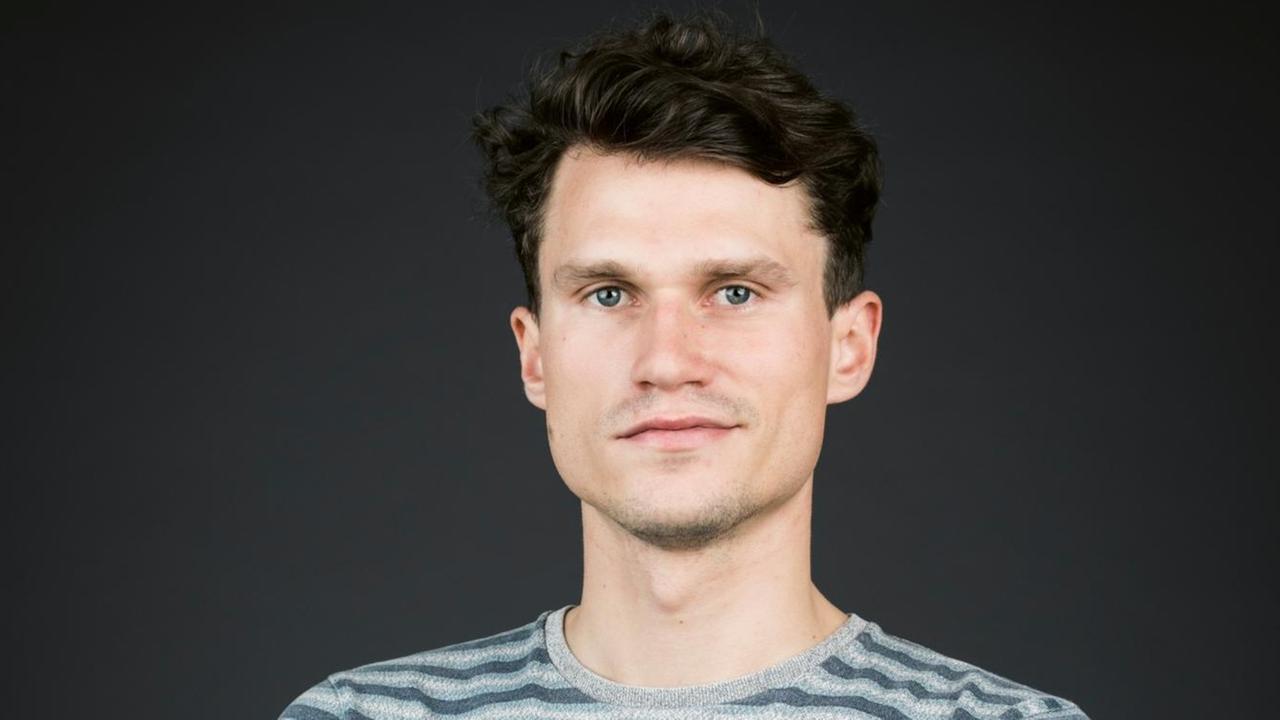 Porträt des Autors Jakob Simmank, im grau gestreiften Pullover vor einem dunklen Hintergrund Porträt des Autors Jakob Simmank, im grau gestreiften Pullover vor einem dunklen Hintergrund