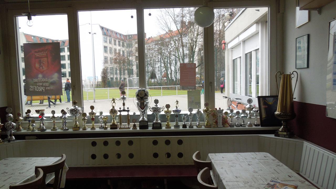 Im Innenraum eines Sport-Vereinsheims stehen Tische und Stühle, auf dem Fensterbrett Pokale. Vor dem Fenster ist ein Sportplatz zu sehen.