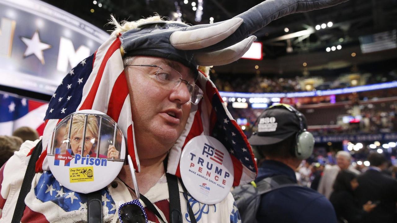 Der Delegierte Michael Lachs mit einer hochgeschobenen Elefantenmaske, einer amerikanischen Flagge um Kopf und Schultern sowie zwei großen Stickern - auf einem steht in Englisch: Hillary Clinton ins Gefängnis, auf dem anderen der Name Trump. Der Delegierte Michael Lachs mit einer hochgeschobenen Elefantenmaske, einer amerikanischen Flagge um Kopf und Schultern sowie zwei großen Stickern - auf einem steht in Englisch: Hillary Clinton ins Gefängnis, auf dem anderen der Name Trump.