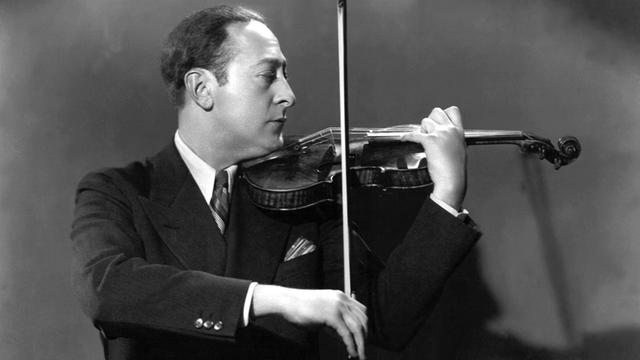 Schwarz-weiß-Fotografie, sie zeigt den Geiger Jascha Heifetz im Profil, er spielt Geige und führt den Bogen mit der rechten Hand. Er hat kurz dunkle Haare, trägt einen schwarzen Anzug mit weißem Hemd und Schlips