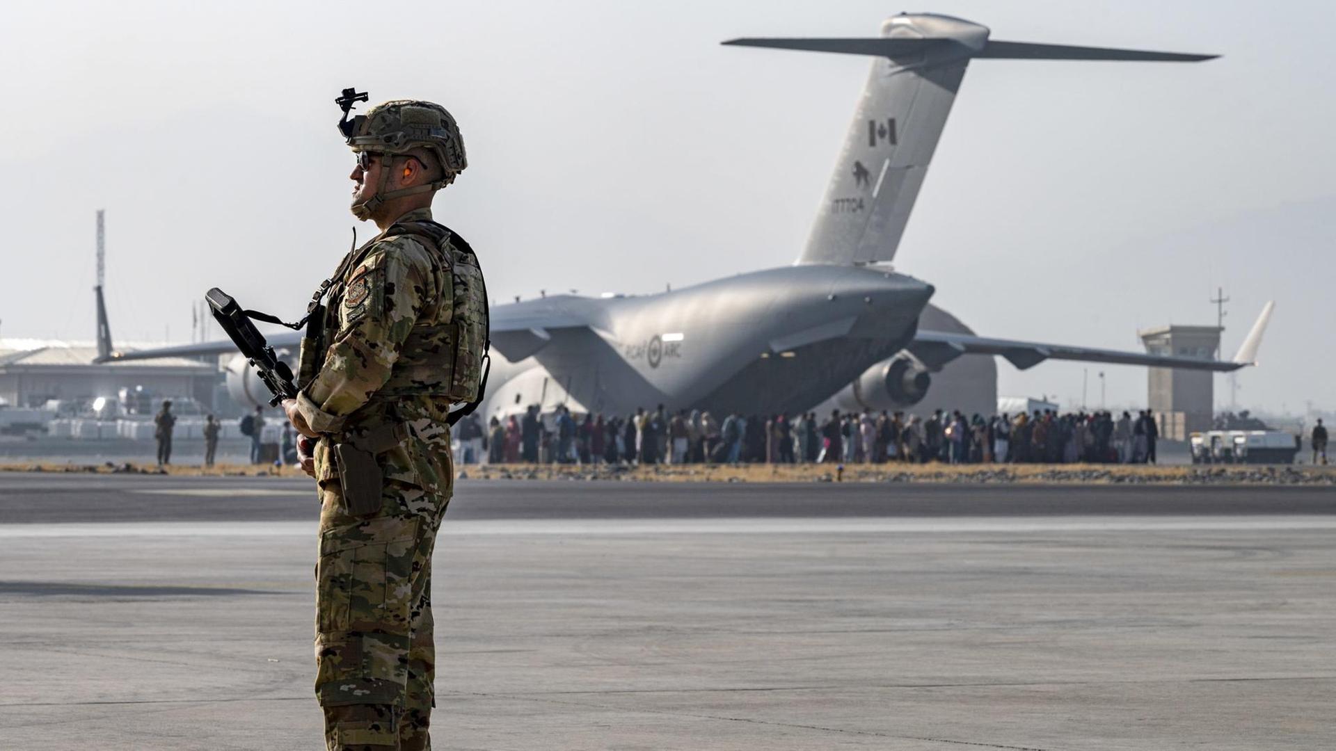 Ein bewaffneter US-Soldat steht am Flughafen Kabul, im Hintergrund ein Flugzeug, das Menschen aus Afghanistan ausfliegen soll. Ein bewaffneter US-Soldat steht am Flughafen Kabul, im Hintergrund ein Flugzeug, das Menschen aus Afghanistan ausfliegen soll.