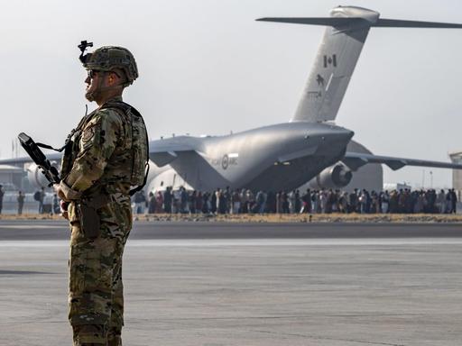 Ein bewaffneter US-Soldat steht am Flughafen Kabul, im Hintergrund ein Flugzeug, das Menschen aus Afghanistan ausfliegen soll.