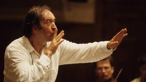 Der Dirigent bei der Arbeit: er fordert die Musiker mit einem Finger vor dem Mund zum leisen Spiel auf.