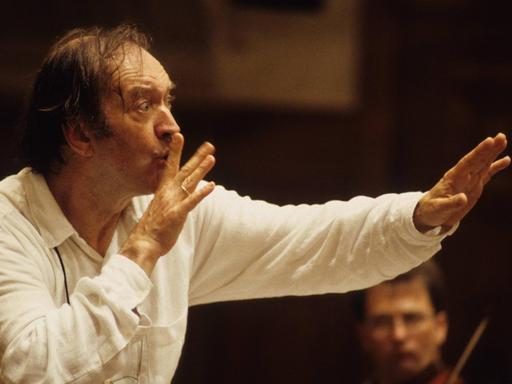 Der Dirigent bei der Arbeit: er fordert die Musiker mit einem Finger vor dem Mund zum leisen Spiel auf.