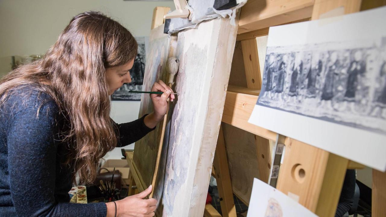 Eine Restauratorin arbeitet an Wandgemälden von der nördlichen Seidenstrasse. Mit einem feinen Pinsel rekonstruiert sie Teile eines aufgebockten Gemäldes. Eine Restauratorin arbeitet an Wandgemälden von der nördlichen Seidenstrasse. Mit einem feinen Pinsel rekonstruiert sie Teile eines aufgebockten Gemäldes.