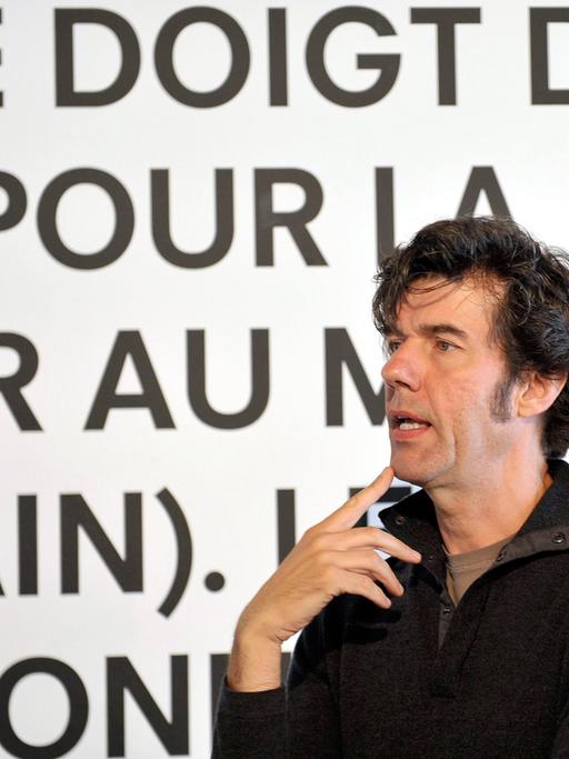 Stefan Sagmeister, schwarzes Hemd, schwarze Haare, steht redend vor einer weißen Wand mit schwarzen, französischen Worten.