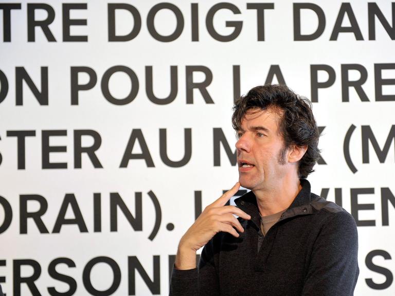 Stefan Sagmeister, schwarzes Hemd, schwarze Haare, steht redend vor einer weißen Wand mit schwarzen, französischen Worten.
