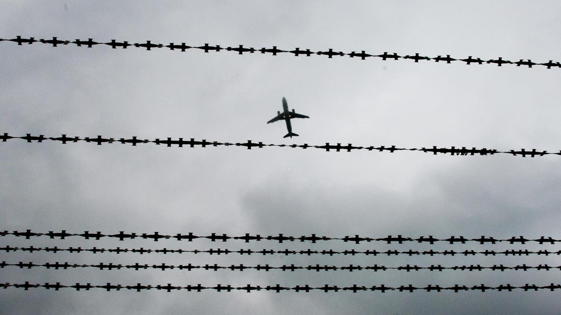 Das Bild zeigt ein Flugzeug am Himmel, im Vordergrund ist Stacheldraht zu sehen. Das Bild zeigt ein Flugzeug am Himmel, im Vordergrund ist Stacheldraht zu sehen.