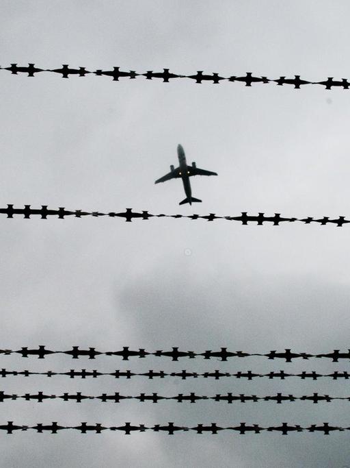 Das Bild zeigt ein Flugzeug am Himmel, im Vordergrund ist Stacheldraht zu sehen.