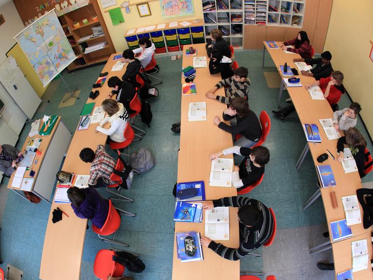 Das Bild ist von oben und mit Fischaugen-Optik fotografiert. Eine Lehrerin unterrichtet Schüler, die Schulbücher aufgeschlagen vor sich liegen haben.