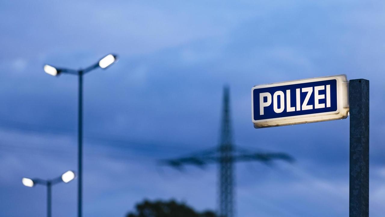 Leuchtschild der Polizeiwache in der Dämmerung. Im Hintergrund sind Straßenlaternen und graue Wolken zu sehen. Leuchtschild der Polizeiwache in der Dämmerung. Im Hintergrund sind Straßenlaternen und graue Wolken zu sehen.