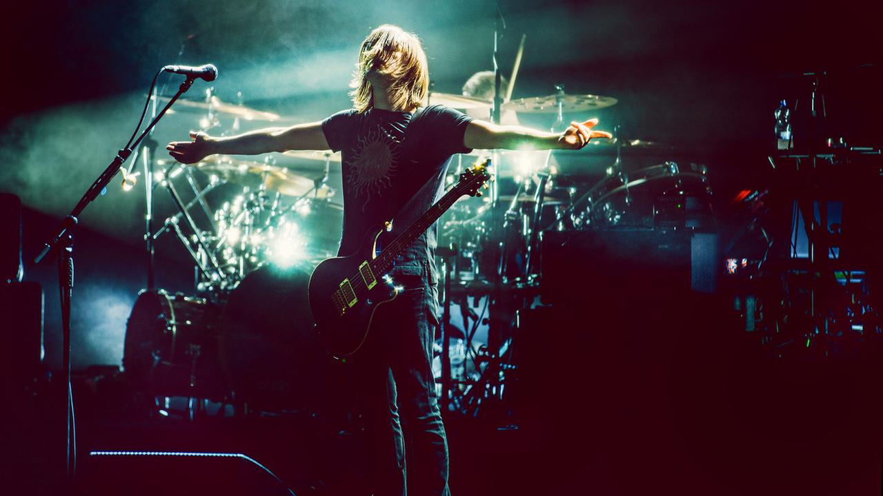 Ein Mann steht auf einer beleuchteten Bühne mit einer umgehängten Gitarre. Er breitet seine Arme aus.