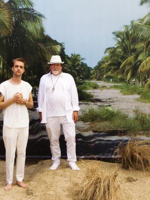 Die Musiker Sebastian und Herbert Janata stehen weißgekleidet in aufgeschüttetem Sand vor einer Fotoleinwan, die einen breiten Weg durch einen Palmenwald zeigt