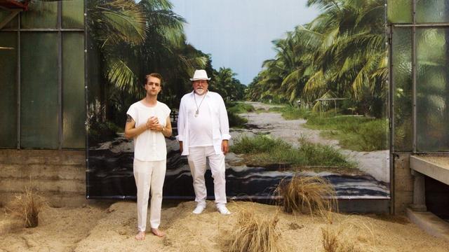 Die Musiker Sebastian und Herbert Janata stehen weißgekleidet in aufgeschüttetem Sand vor einer Fotoleinwan, die einen breiten Weg durch einen Palmenwald zeigt