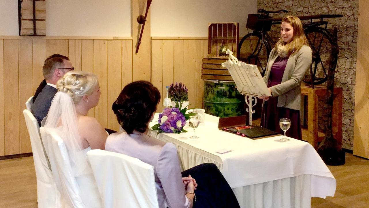 Ein Hochzeitspaar sitzt an einem Tisch, eine Rednerin steht dahinter. Ein Hochzeitspaar sitzt an einem Tisch, eine Rednerin steht dahinter.