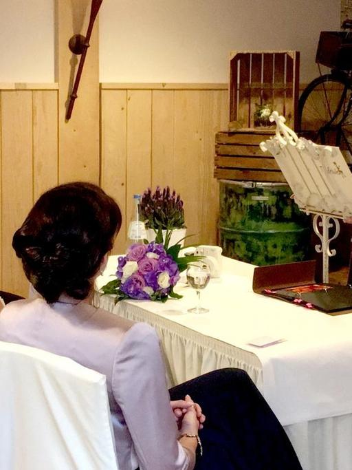 Ein Hochzeitspaar sitzt an einem Tisch, eine Rednerin steht dahinter.