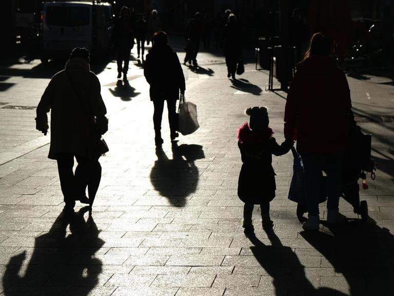 Passanten mit Tüten gehen während des Lockdowns durch eine Fußgängerzone und sind dabei als Silhouette und Schatten zu sehen.