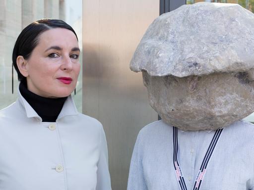 Das Bild zeigt die Künstlerin Natascha Süder Happelmann mit einer Steinskulptur auf dem Kopf, die ihr Gesicht verbirgt, und ihre Sprecherin Helene Duldung.