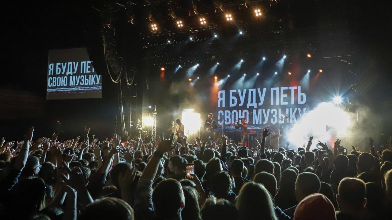 Im Hintergrund eine Bühne mit russischer Schrift. Davor eine Zuschauermenge.