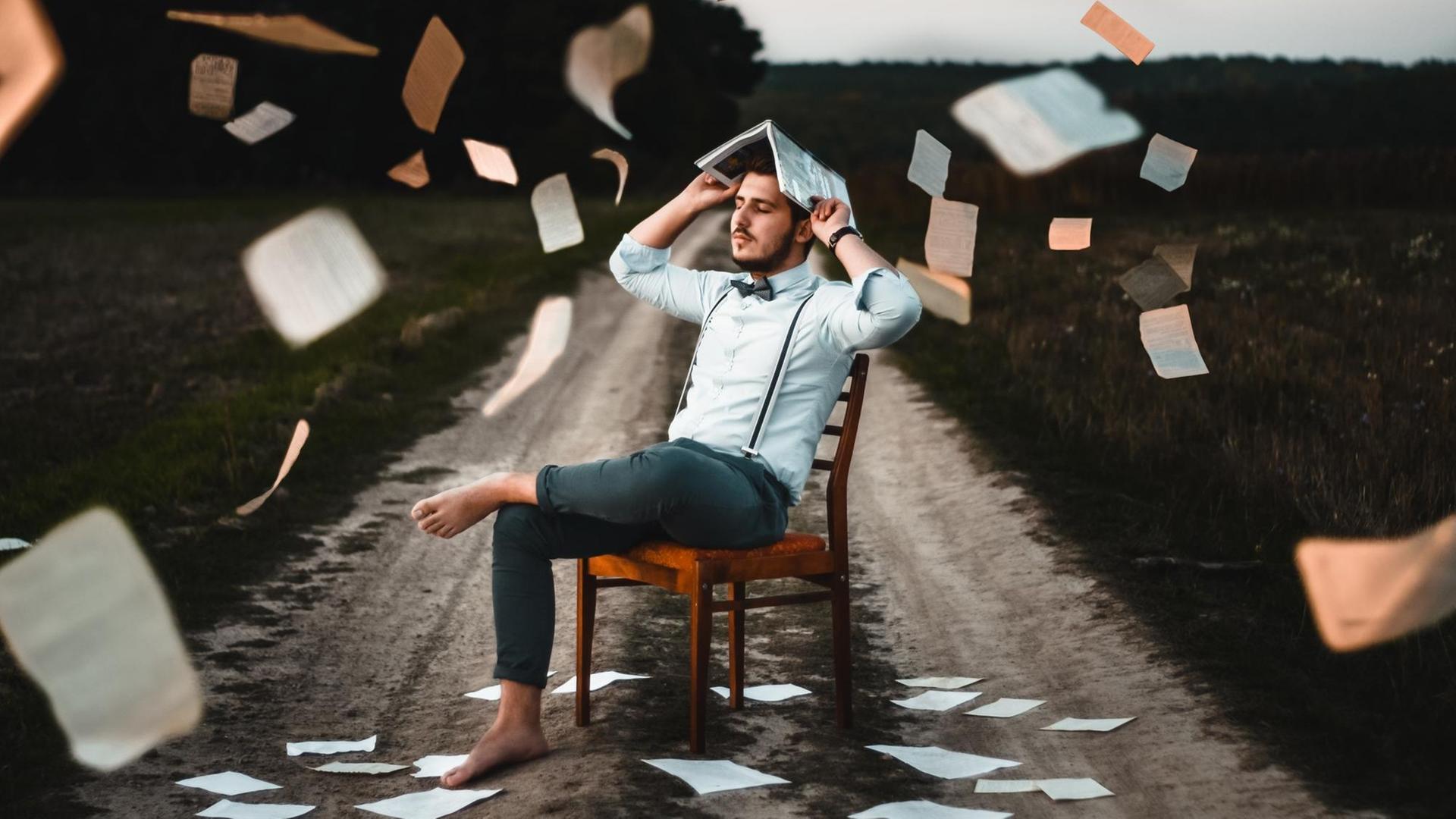 Das Bild zeigt einen Mann in Hemd, mit Hosenträgern und Fliege. Er sitzt auf einem Stuhl, draußen auf einem Feldweg. Er hält sich ein Magazin schützend über den Kopf, während es Papierseiten von oben regnet. Das Bild zeigt einen Mann in Hemd, mit Hosenträgern und Fliege. Er sitzt auf einem Stuhl, draußen auf einem Feldweg. Er hält sich ein Magazin schützend über den Kopf, während es Papierseiten von oben regnet.