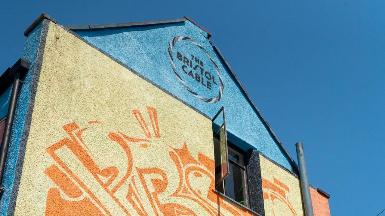 Die Seitenwand eines Gebäudes, das blau-gelb gestrichen und mit orangefarbenen Mustern bemalt ist. Am Giebel steht die Aufschrift "The Bristol Cable".