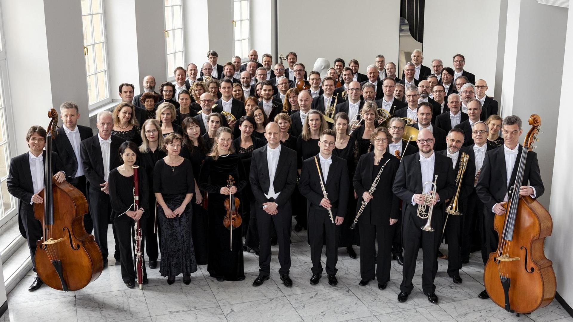 Das Orchester steht in Abendrobe und Instrumenten im hellen Foyer des Opernhauses und schaut leicht nach oben in die Kamera. Das Orchester steht in Abendrobe und Instrumenten im hellen Foyer des Opernhauses und schaut leicht nach oben in die Kamera.