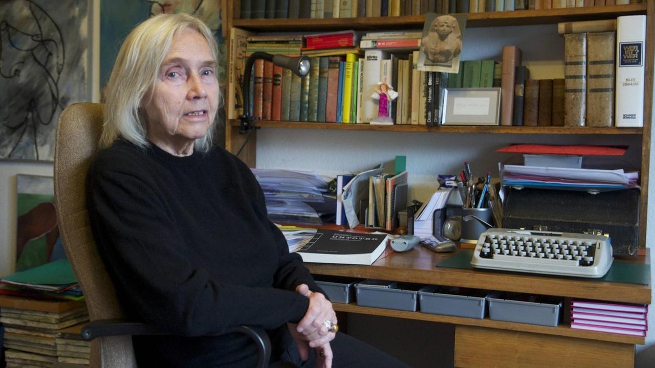 Eine ältere Frau sitzt an einem Schreibtisch vor einer Bücherwand. An den Wänden sind Malereien zu sehen. Eine ältere Frau sitzt an einem Schreibtisch vor einer Bücherwand. An den Wänden sind Malereien zu sehen.
