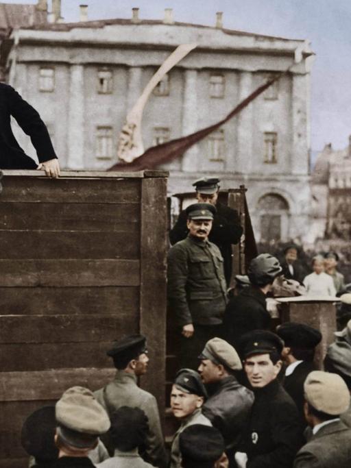 Wladimir Iljitsch Lenin auf einem Podest, er spricht a, 5. Mai 1920 auf dem Swerdlow-Platz (ehem. Theaterplatz) in Moskau zu Soldaten der Roten Armee vor deren Abmarsch an die Front im Russisch-Polnischen Krieg; rechts auf den Stufen der Tribüne stehend: Leo Trotzkij (vorn) und Lew Kamenew (digital koloriert)