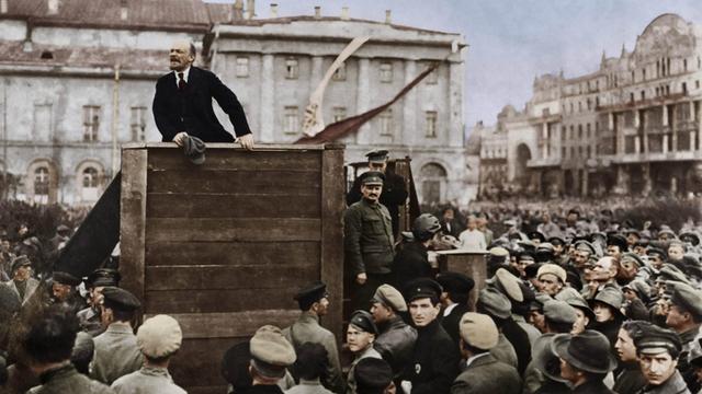 Wladimir Iljitsch Lenin auf einem Podest, er spricht a, 5. Mai 1920 auf dem Swerdlow-Platz (ehem. Theaterplatz) in Moskau zu Soldaten der Roten Armee vor deren Abmarsch an die Front im Russisch-Polnischen Krieg; rechts auf den Stufen der Tribüne stehend: Leo Trotzkij (vorn) und Lew Kamenew (digital koloriert)