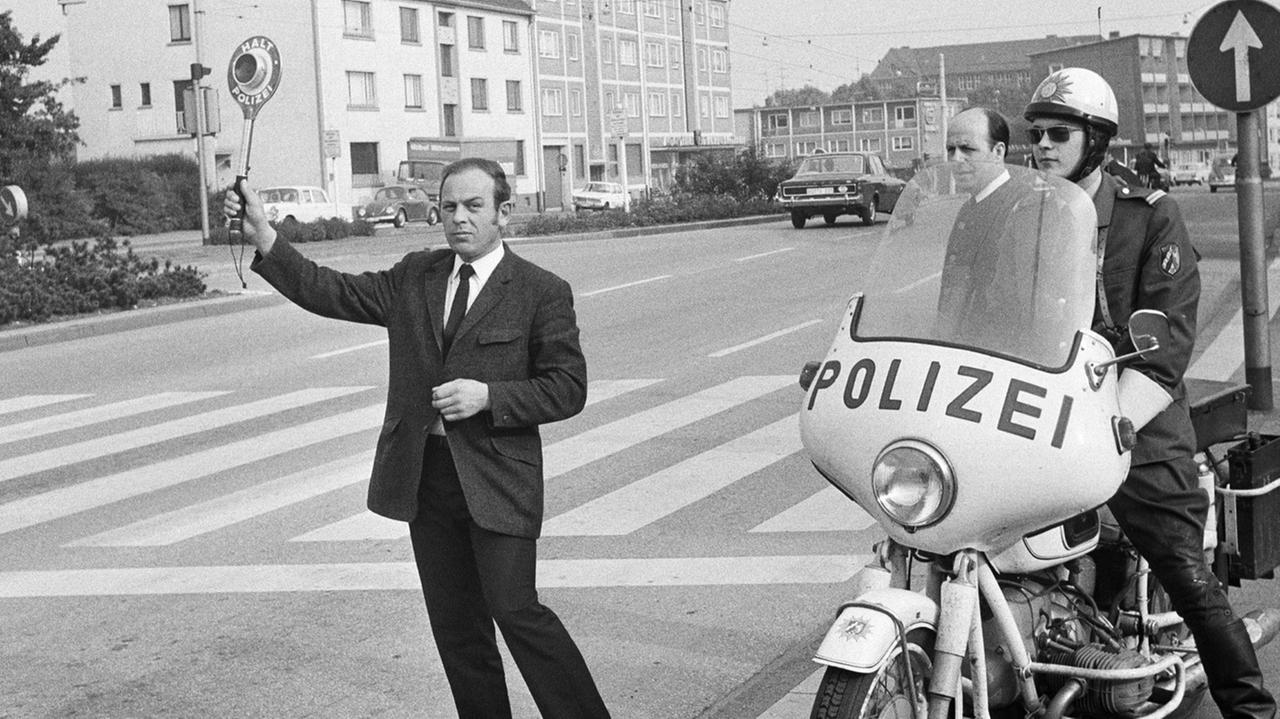 Verkehrskontrolle 1970 in Bottrop. An einer Straße steht ein Polizist mit einem Motorrad. Ein anderer winkt mit einer Polizeikelle Verkehrsteilnehmer raus.