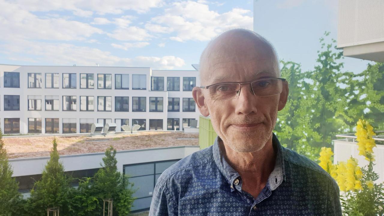 Ein älterer Herr mit Brille steht auf einem Balkon und schaut milde lächelnd in die Kamera - hinter ihm sind der Himmel und ein Spielplatz zu erkennen. Ein älterer Herr mit Brille steht auf einem Balkon und schaut milde lächelnd in die Kamera - hinter ihm sind der Himmel und ein Spielplatz zu erkennen.