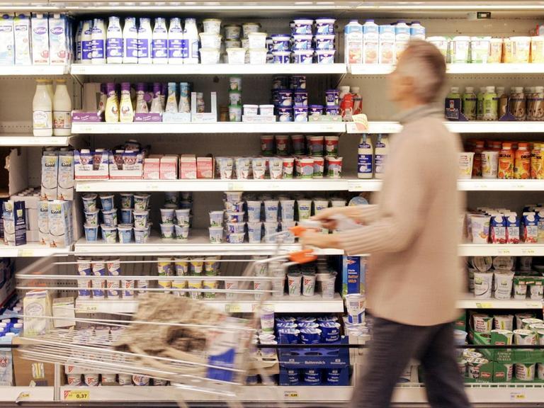 Ein Supermarktregal mit Plastikverpackungen. Davor geht ein grauhaariger Mann mit einem Einkaufswagen.