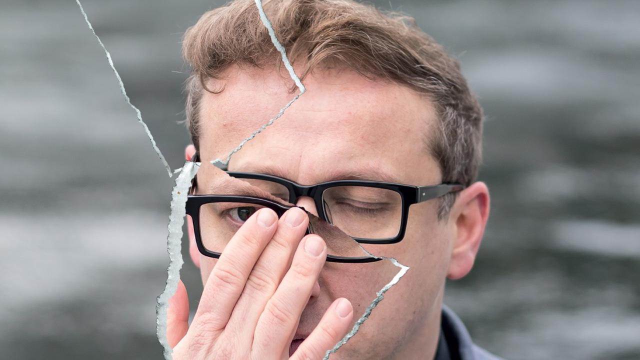 Ein Mann mit schwarzer Brille schaut durch eine zerbrochene Scheibe. Er hält die rechte Hand vor sein Gesicht. Ein Mann mit schwarzer Brille schaut durch eine zerbrochene Scheibe. Er hält die rechte Hand vor sein Gesicht.