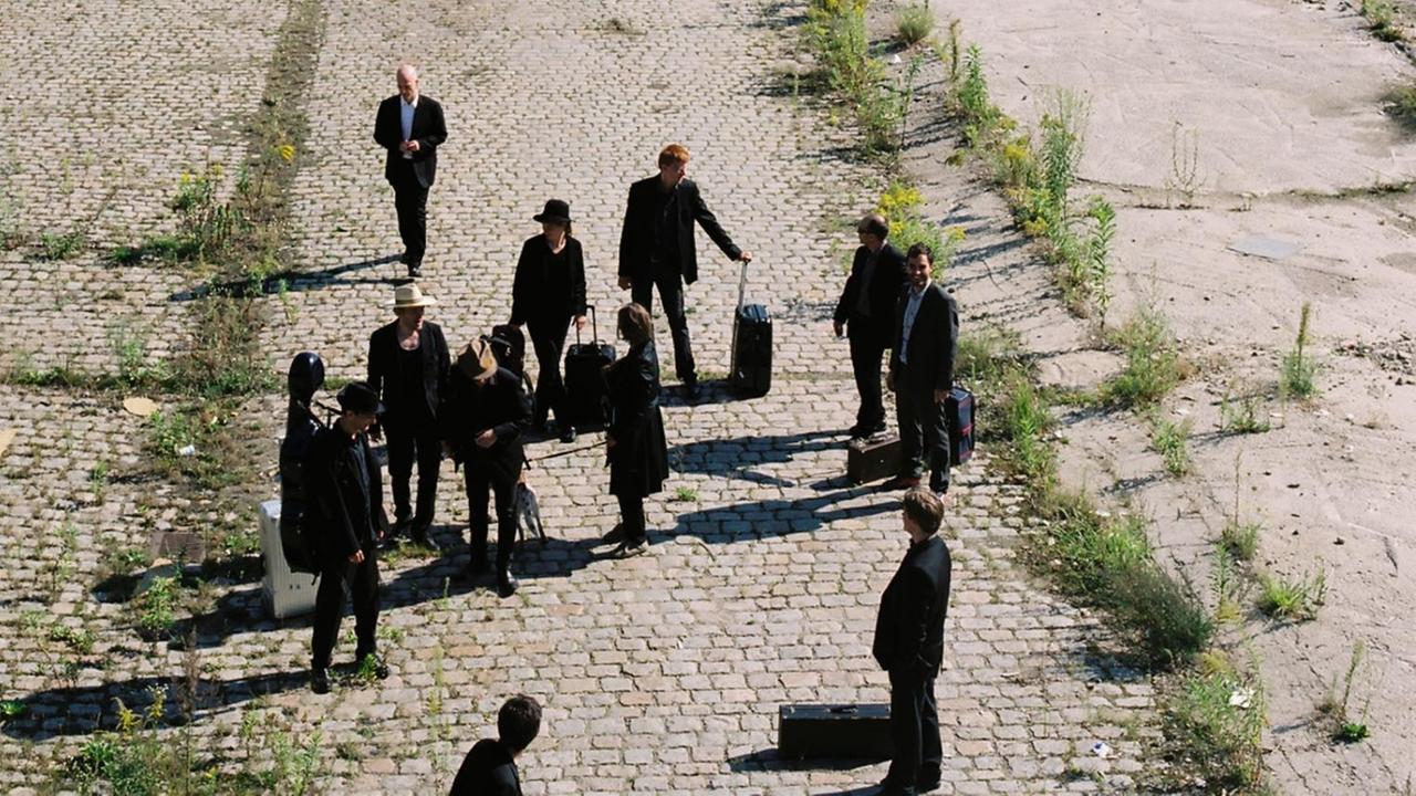Eine Gruppe von Menschen steht mit Reisekoffern auf einem verlassenen Bahnhofsgelände. Eine Gruppe von Menschen steht mit Reisekoffern auf einem verlassenen Bahnhofsgelände.