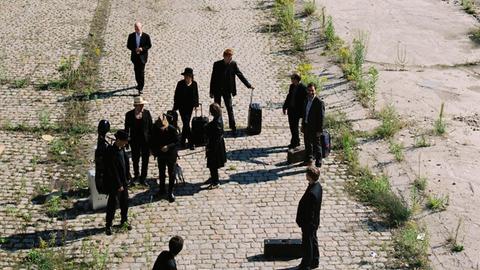 Eine Gruppe von Menschen steht mit Reisekoffern auf einem verlassenen Bahnhofsgelände.