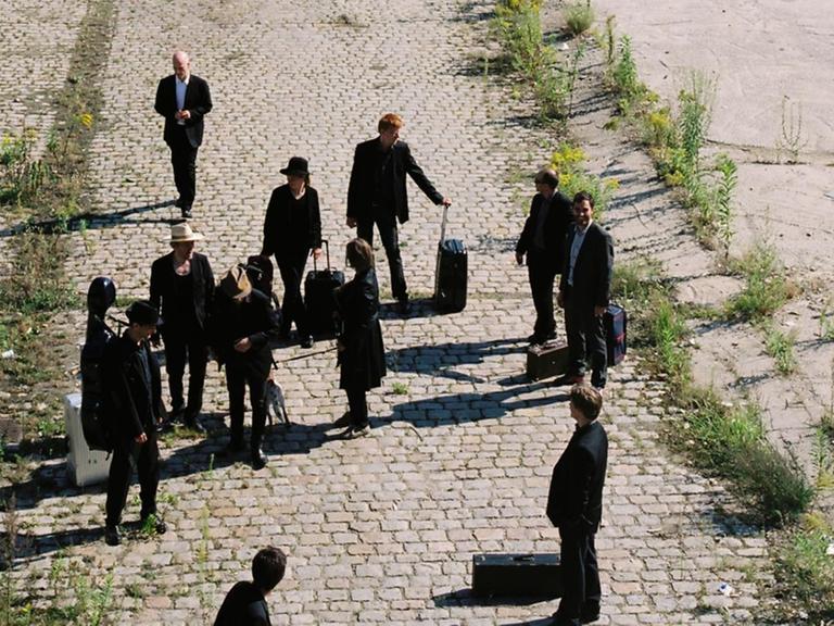Eine Gruppe von Menschen steht mit Reisekoffern auf einem verlassenen Bahnhofsgelände.