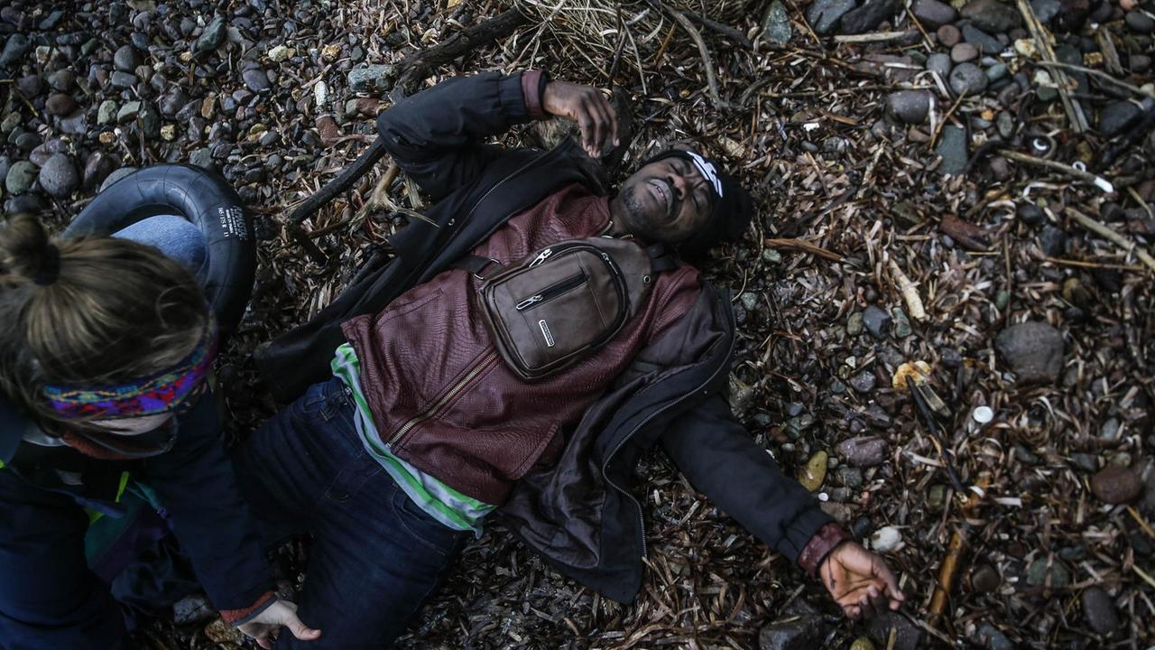 Ein Mann liegt erschöpft am Steinstrand von Lesbos während eine Frau sein linkes Bein abtastet. Ein Mann liegt erschöpft am Steinstrand von Lesbos während eine Frau sein linkes Bein abtastet.